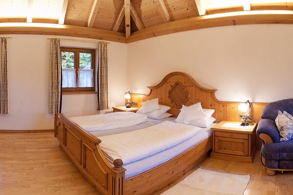 Schlafzimmer der Ferienwohnung Vorderhorn mit Holz-Doppelbett, Nachtkästchen und blauem Sessel unter der Holzdecke